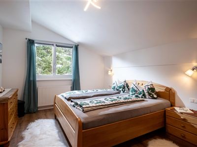 Ein gemütliches Schlafzimmer mit einem Doppelbett und Holzmobiliar. Große Fenster lassen viel Licht herein und bieten einen Blick ins Grüne.