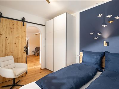 Een moderne slaapkamer met een groot bed en een elegante witte commode. Aan de muur hangen decoratieve vogels en een comfortabele stoel staat klaar.