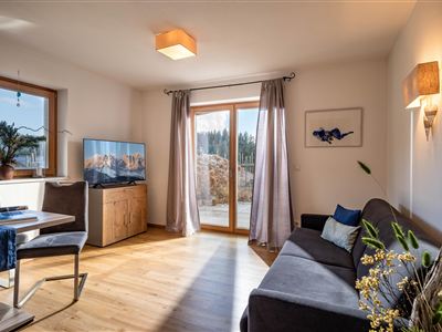 A cozy living room with a sofa, a dining table, and a television. Generous windows let in plenty of light and offer a view of the outdoors.