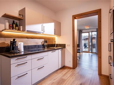 A modern kitchen with white cabinets and a dark countertop. An open passageway reveals the living area, which is illuminated by natural light.