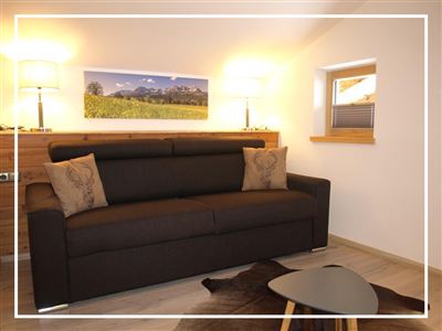 Ein gemütliches Wohnzimmer mit einem dunkelbraunen Sofa und dekorativen Kissen. Die Wände sind hell und es gibt einen kleinen Tisch sowie eine Fensteröffnung mit natürlichem Licht.