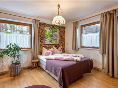 A cozy bedroom with wooden flooring and a large bed. The walls are decorated, and there are windows with curtains as well as a plant in the corner.
