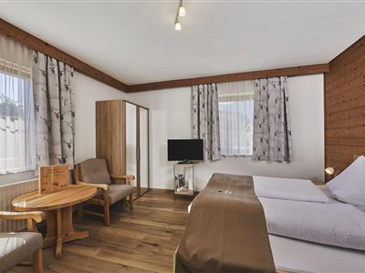A cozy room with wooden paneling and two beds. There is a table, chairs, and a television, as well as plenty of daylight coming through the windows.