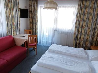 Ein gemütliches Hotelzimmer mit zwei Betten und einem roten Sofa. Große Fenster lassen viel Licht herein, und der Raum ist einfach eingerichtet.