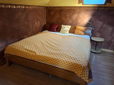 Een gezellige slaapkamer met een groot bed, dat is versierd met een oranje deken en kleurrijke kussens. De muren zijn in warme aardetinten gehouden en er staat een kleine nachttafel naast het bed.