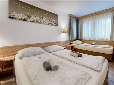 A cozy triple room with multiple beds and wooden paneling. Large windows let in plenty of daylight.