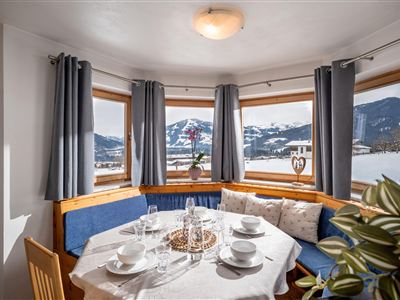 Eine gemütliche Essecke mit einer runden Tischgruppe und einer gepolsterten Sitzbank. Die Fenster sind mit Vorhängen geschmückt, und der Raum wird von einer Deckenleuchte beleuchtet.