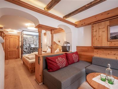 A cozy room with wooden interiors and a modern seating area. In the background, a bedroom with an appealing wall design is visible.