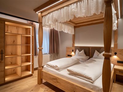 A cozy bedroom with a wooden four-poster bed and white bed linens. In the background, there is a wooden shelf.