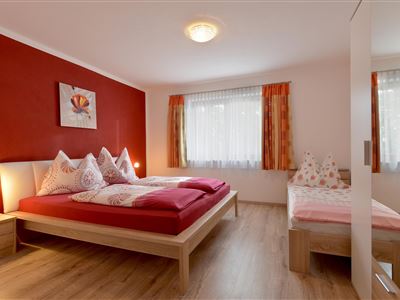 A cozy bedroom with a large double bed and a single bed. The walls are painted red and there are large windows with curtains.