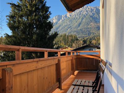 A balcony with a wooden railing and two chairs. In the background, there are high mountains and a clear blue sky.