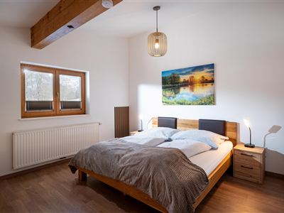Een gezellige slaapkamer met een groot bed en een warme houten plafond. Aan de muur hangt een afbeelding van een landschap, en het licht komt binnen via twee vensters.