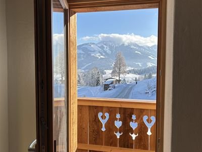 Ein offenes Fenster mit Blick auf einen verschneiten Balkon und die Berge im Hintergrund. Die Sonne scheint auf die winterliche Landschaft.