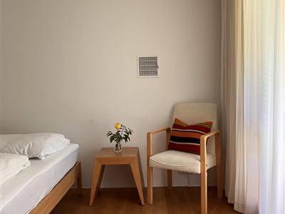 Ein gemütliches Schlafzimmer mit einem Bett und einem Stuhl. Ein kleiner Tisch mit einer Blume und ein bunter Teppich ergänzen den Raum.