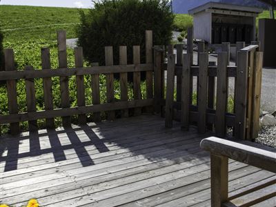 Een houten terras met een klein hek en een bloempot met gele bloemen. Op de achtergrond zijn groene weides en een paar bergen zichtbaar.