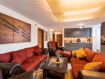A cozy living room with red sofas and an elegant table. The kitchen in the background is modern and well-equipped.