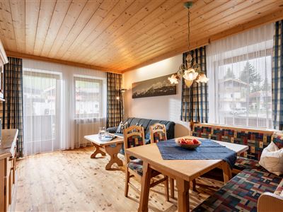 A cozy living space with a wooden ceiling and large windows. There is a dining table, a sofa, and a decorative lamp.