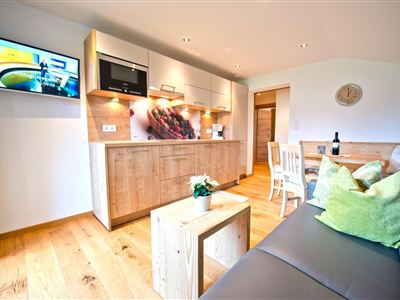 Eine moderne Küche mit Holzregalen und einem eleganten Esstisch. Im Hintergrund ist ein Fernseher und gemütliche Sitzmöglichkeiten zu sehen.