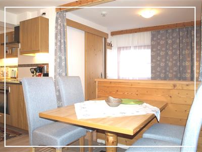A cozy kitchen nook with a wooden table and chairs. In the background, modern kitchen cabinets and curtains can be seen.