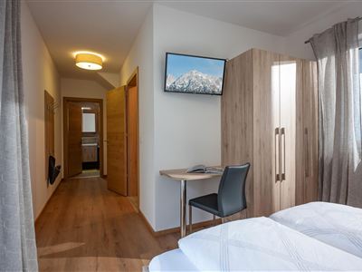 A modern room with a double bed and a table. A television hangs on the wall, and there is a view of a hallway with wooden details.