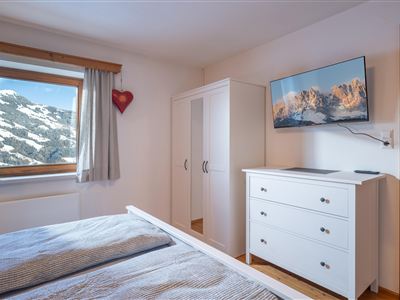 Ein gemütliches Schlafzimmer mit einem Doppelbett, einem Kleiderschrank und einem Fernseher an der Wand. Durch das Fenster ist eine schneebedeckte Berglandschaft zu sehen.