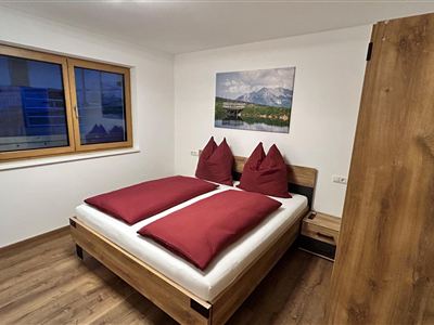 Een gezellige slaapkamer met een tweepersoonsbed en rode kussens. De muren zijn licht, en een raam laat daglicht binnen.