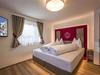 Ein modernes Schlafzimmer mit einem Doppelbett und einem großen Fernseher. Die Wände sind hell, und es gibt große Fenster mit Vorhängen.