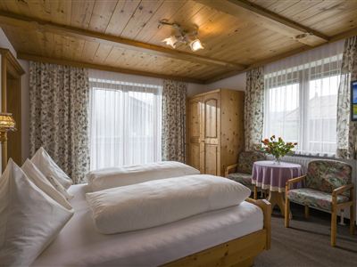 Ein gemütliches Zimmer mit zwei Betten, Holzdecken und hellen Vorhängen. Es gibt einen kleinen Tisch mit Blumen und einen Fernseher an der Wand.