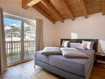 Ein gemütliches Schlafzimmer mit Holzbalkendecke und einem großen Fenster. Das Bett ist mit Kissen und Decken dekoriert und bietet einen Blick auf die Berge.
