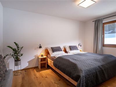 Een moderne slaapkamer met een groot bed en gezellige beddengoed. De ruimte heeft een raam en is in lichte kleuren gehouden, met een plantenbak in de hoek.