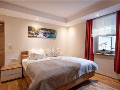 Ein gemütliches Schlafzimmer mit einem großen Bett und weichen Kissen. Die Wände sind hell, und es gibt Vorhänge in einem warmen Farbton sowie ein Fenster mit Pflanzen.