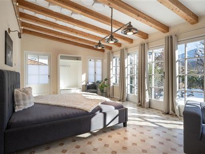 A bright, modern bedroom with a wooden beam ceiling and large windows. The bed is cozy, decorated with a blanket and pillows.