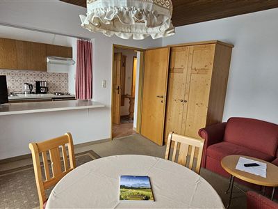 Ein gemütlicher Wohnraum mit einem Tisch, Stühlen und einem roten Sofa. Im Hintergrund sieht man eine Küchenzeile und einen Schrank.