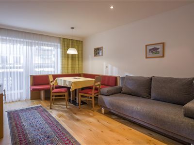 Ein gemütlicher Wohnbereich mit einem grauen Sofa und einem Esstisch. Große Fenster sorgen für viel Tageslicht.