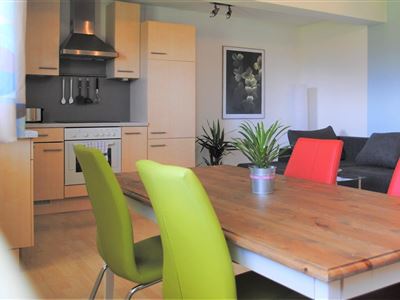 A modern kitchen with wooden cabinets and a large dining table. The chairs are in vibrant colors, and there are plants in the corner.