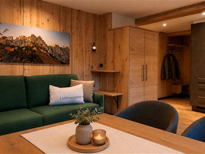 Ein modernes Wohnambiente mit Holzelementen und einem gemütlichen Sofa. Im Hintergrund ist eine Küche sichtbar, und ein Bild von bergiger Landschaft ziert die Wand.