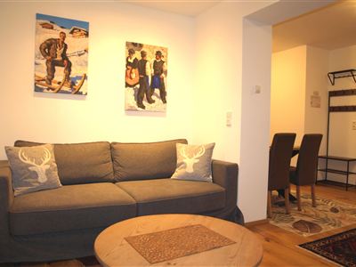 Ein gemütliches Wohnzimmer mit einem grauen Sofa und dekorativen Kissen. An der Wand hängen Bilder und ein Essbereich mit Stühlen ist sichtbar.