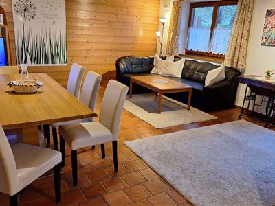 A cozy living space with a wooden ceiling, a dining table, and elegant white chairs. A couch and decorative elements create an inviting atmosphere.
