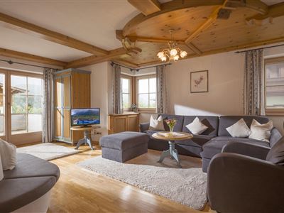 A cozy living room with a large sofa, a wooden table, and a television. Large windows let in plenty of light and offer a view outside.