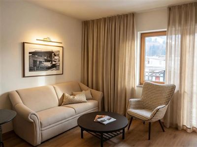 A cozy living room with a beige sofa and an upholstered armchair. Large windows and curtains provide plenty of natural light.