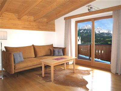 A cozy living room with a brown sofa and a table. The window offers a view of the mountains and nature.