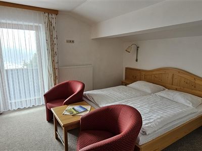 Ein gemütliches Hotelzimmer mit einem Doppelbett und zwei roten Sessel. Große Fenstertüren sorgen für viel Tageslicht.