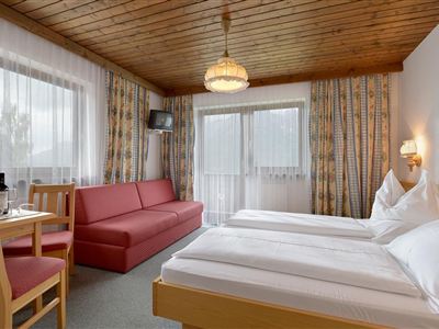 A cozy hotel room with two single beds and a red sofa. The room has large windows and an inviting atmosphere.