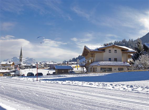 Eine malerische Winterlandschaft mit schneebedeckten Häusern und Bergen. Der Himmel ist klar mit einigen Wolken und ein Gleitschirm fliegt darüber.