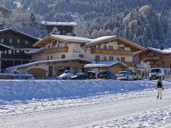 Ein malerisches Chalet im Schnee mit einem klaren blauen Himmel. Im Vordergrund läuft eine Person auf einem verschneiten Weg.