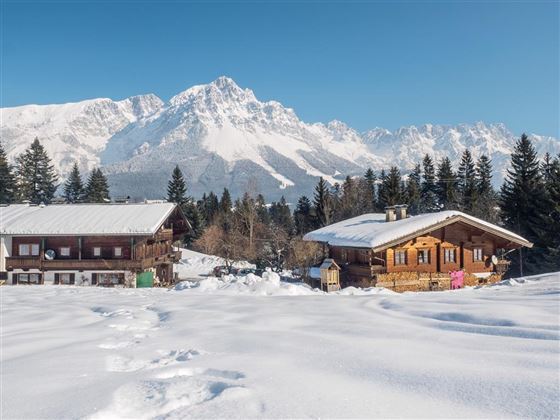 An idyllic winter scene with snow-covered cottages and majestic mountains in the background. The clear blue sky completes the peaceful atmosphere.
