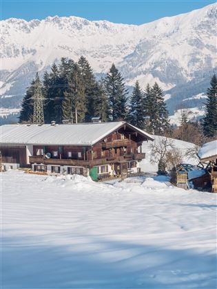 A picturesque chalet in a snow-covered landscape with mountains in the background. The surroundings are tranquil and surrounded by coniferous trees.