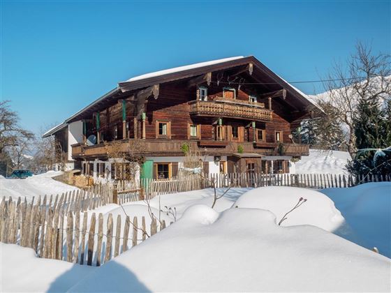A traditional wooden house in the snow with a clear blue sky. The surroundings are covered by a white blanket of snow and a simple wooden fence.