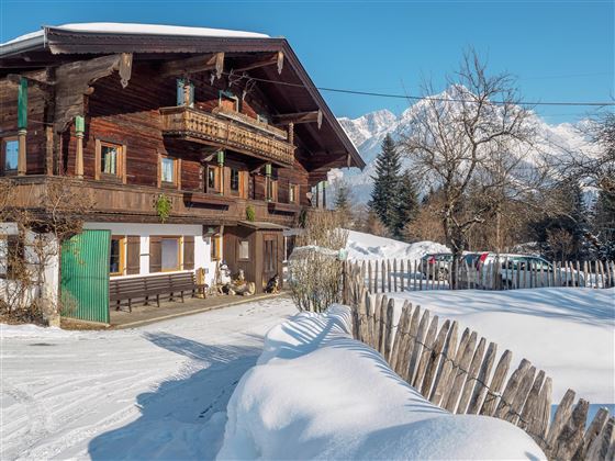 A traditional wooden house in the snow with a clear blue sky in the background. The landscape is surrounded by snow-covered trees.