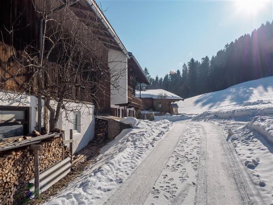 A snowy path leads alongside traditional wooden houses. The surroundings are quiet and surrounded by snow-covered mountains.